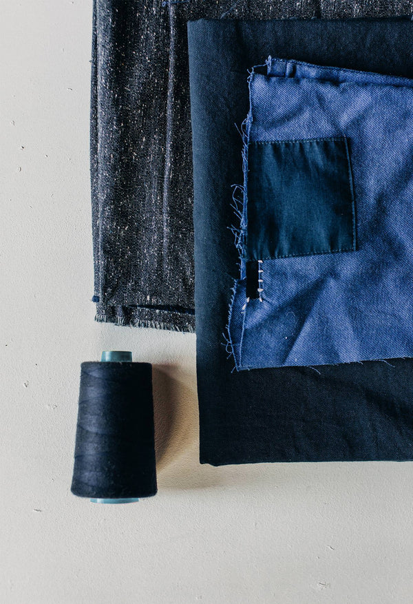 Swatches of blue fabrics are folded atop a white surface near a spool of black thread.