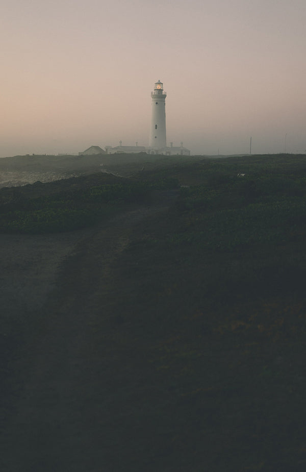 A lighthouse and connected buildings across a foggy shoreline at dusk.