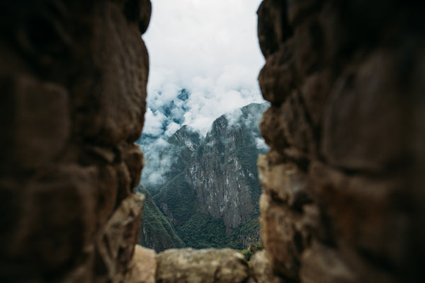 Looking through a slotted window in a stone wall, out and down over clouded mountain peaks.